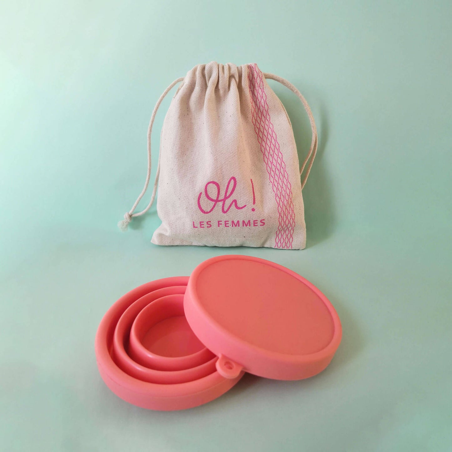 Stérilisateur à disque ou cup en coloris corail plié et ouvert posé avec son pochon en coton imprimé logo Oh les femmes le tout sur fond vert d'eau