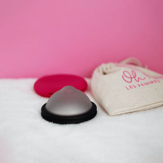 Disque menstruel discoh taille L coloris noir avec au fond sa pochette en silicone de rangement et à sa droite le pochon en coton imprimé le tout sur fond rose
